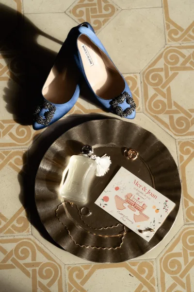 Wedding details flatlay with wedding invitation flatlay, blue embellished heels, perfume, gold rings and necklace on a metal tray over sunlit tile floor