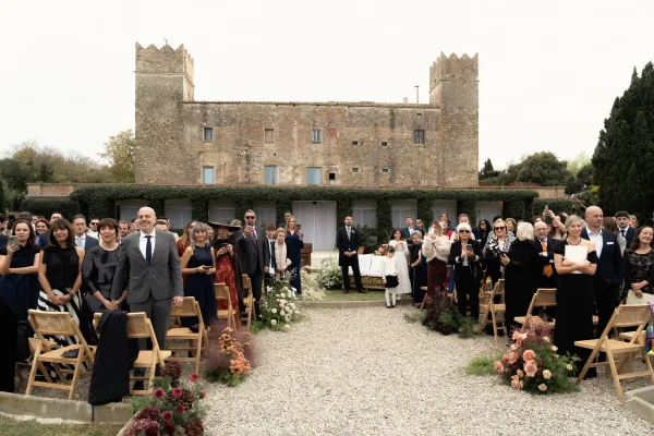 Ceremony processional at an outdoor wedding ceremony, guests standing by a gravel aisle lined with floral arrangements in a castle courtyard