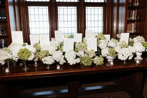 Seating chart display on an escort card table accented with hydrangeas, set before leaded windows, with bookshelves in the background.