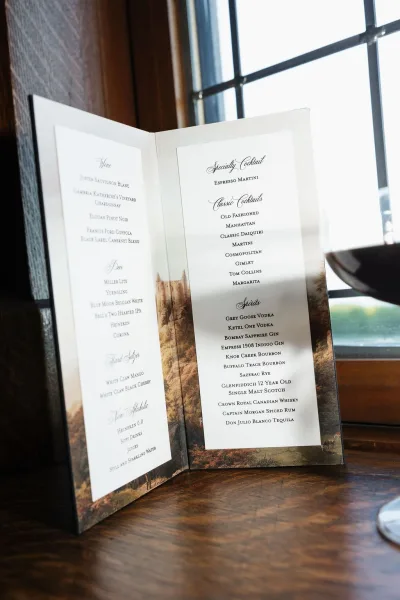 Wedding drink menu presented as a bar menu sign on a wood tabletop by window panes, printed with elegant calligraphy script.