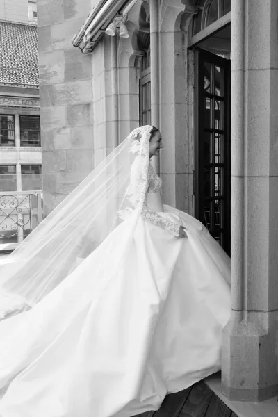 Bridal portrait of a bride with long veil, her cathedral veil sweeping across a stone balcony before arched windows and distant cityscape.