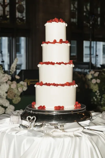 Wedding cake, a four tier cake with smooth white icing crowned by fresh raspberries, set before tall windows with city buildings beyond.