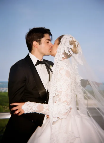 Wedding kiss portrait as a bride and groom kiss beneath a lace veil, framed by blue sky, distant trees, and a shimmering water view.