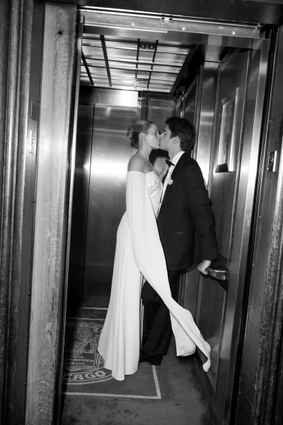 Wedding kiss portrait as the bride and groom kiss inside an elevator with metal doors and wood paneling, the bride’s cape sleeves framing them.
