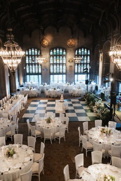 Reception setup for an indoor wedding reception with low floral centerpieces beneath stained glass windows and a vaulted wood ceiling.