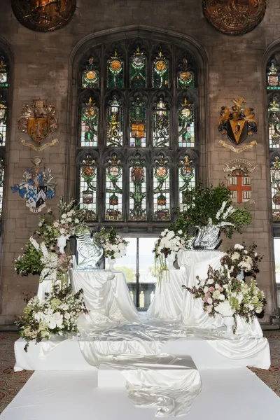Ceremony setup for elegant indoor ceremony decor with white satin draping, framed by stained glass windows and a stone wall with heraldic crests.