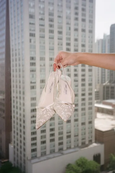 Bridal shoes styled as elegant slingback wedding shoes with pearl detailing, held by a window overlooking the city skyline and trees.