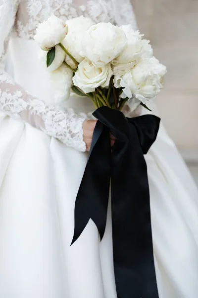 Bridal bouquet, a white peony bouquet finished with a black satin ribbon, held against a weathered stone wall.