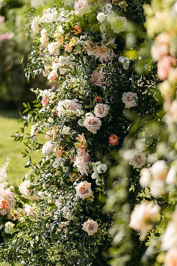 Wedding floral arch with blush and white flowers, roses and dahlias, accented by greenery on a sunlit garden lawn with trees behind