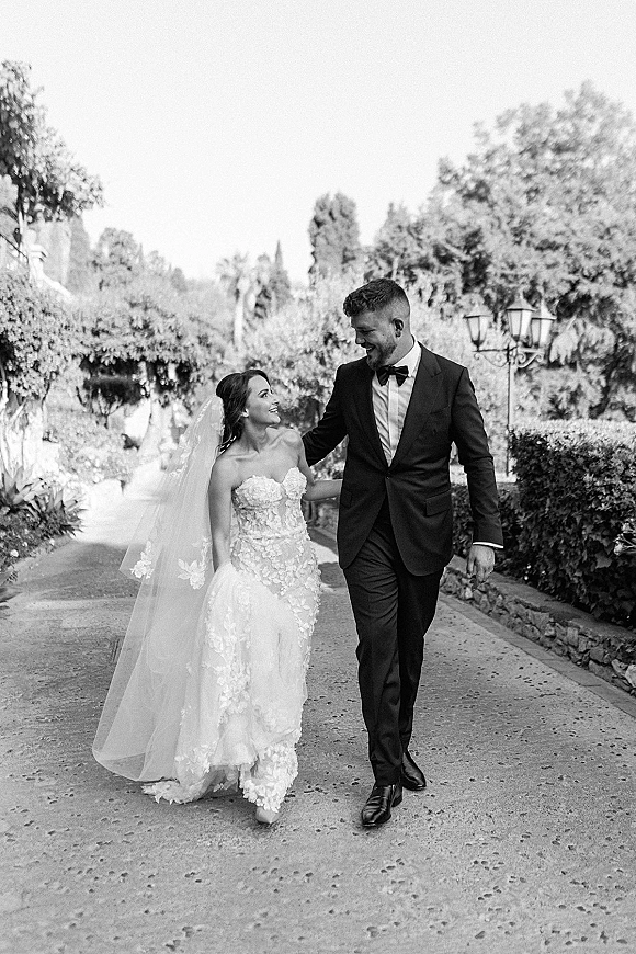 Couple portrait in a black and white wedding portrait style, bride in strapless lace gown and long veil walking with groom in tuxedo on a tree-lined garden path