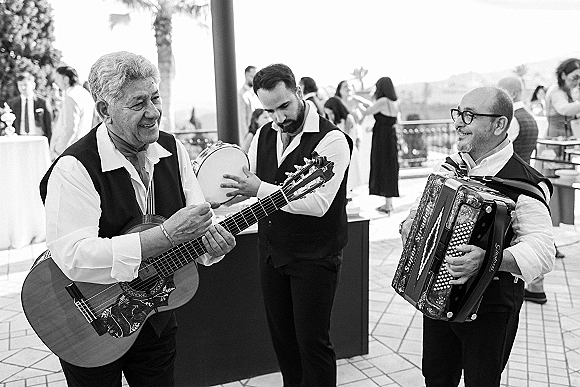 Wedding live band of wedding musicians playing acoustic guitar, accordion, and tambourine on an outdoor terrace with palm trees behind guests