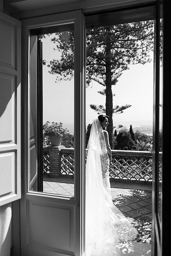 Bridal portrait in a lace gown with long veil and train, standing on a balcony terrace framed by open French doors, looking away