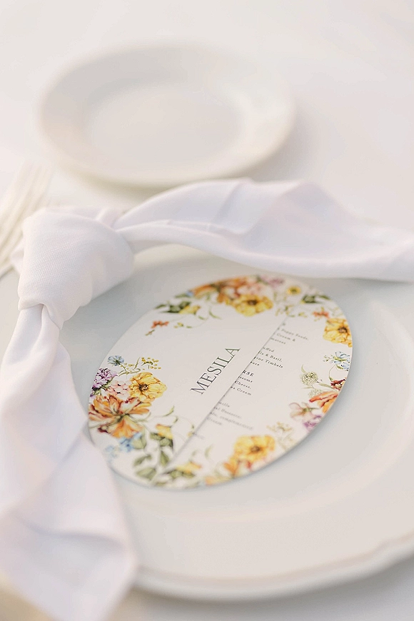 Wedding menu card with floral accents on a white napkin over a dinner plate, styled in an oval wedding menu design on a white table setting