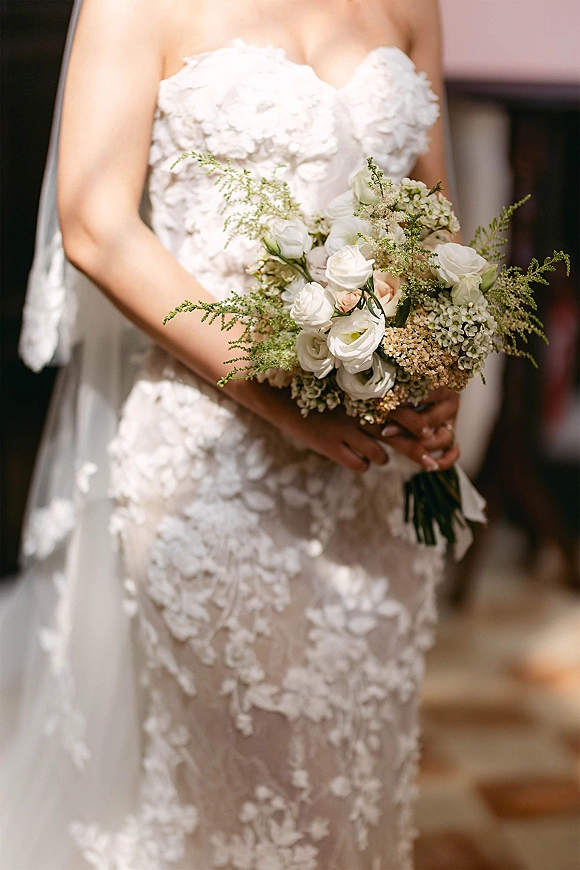 Bridal bouquet of white and blush roses with greenery, held by a bride in a strapless lace dress and veil in a softly blurred hallway