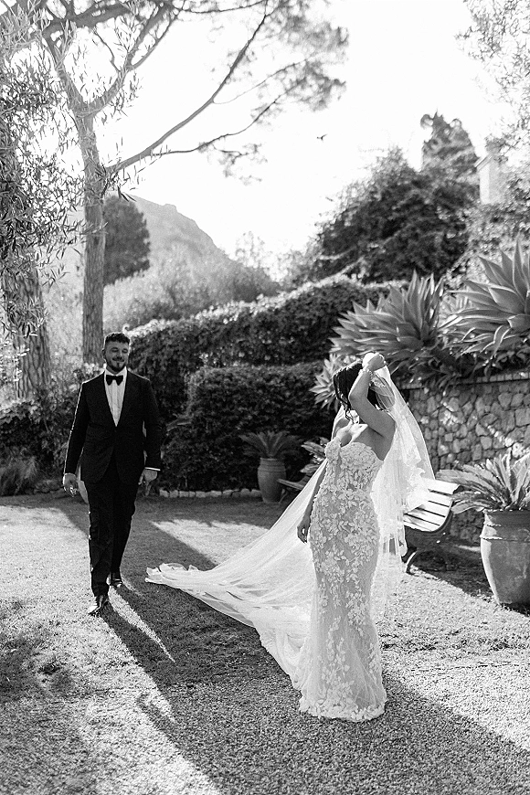 First look moment as bride adjusts her long wedding veil while groom in black tuxedo walks toward her in a garden by a stone wall