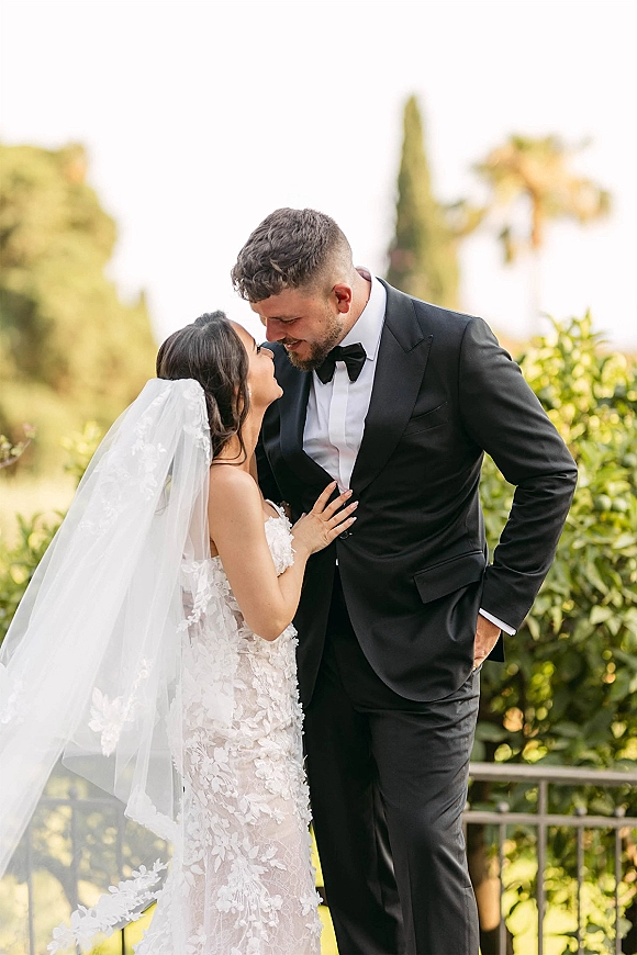 Couple portrait of bride and groom portrait touching foreheads, her lace dress and veil flowing as she holds his tuxedo lapel outdoors