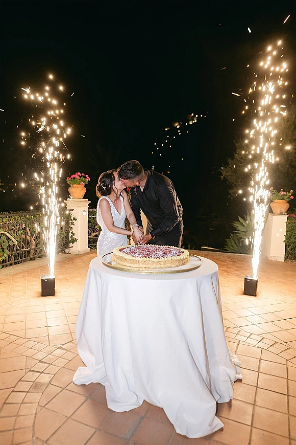 Wedding cake cutting as bride and groom slice the cake on a round table, cold spark fountains behind them on an outdoor patio at night