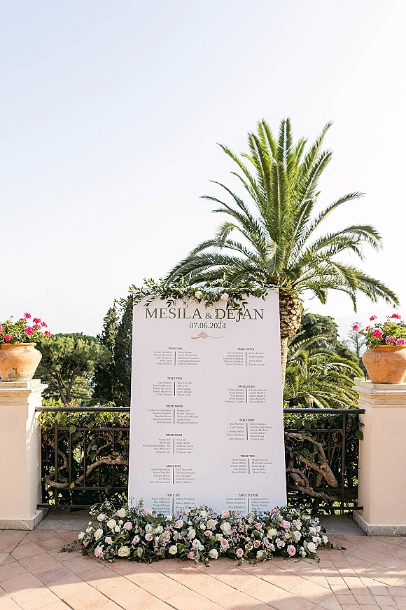 Seating chart sign with calligraphy wedding seating chart, greenery garland and blush roses on a terrace patio with palm tree backdrop