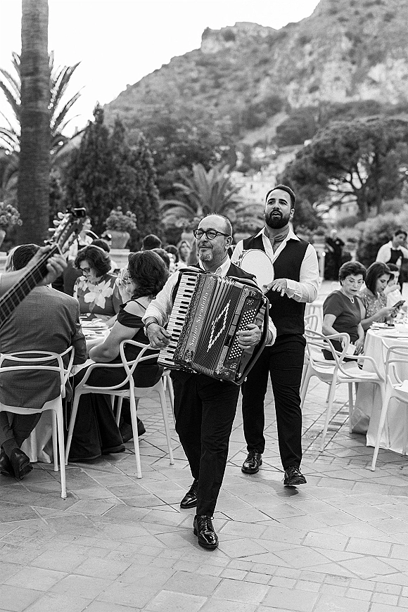 Wedding reception band playing live wedding music with an accordion and tambourine, walking past banquet tables on an outdoor terrace with palm trees and mountains
