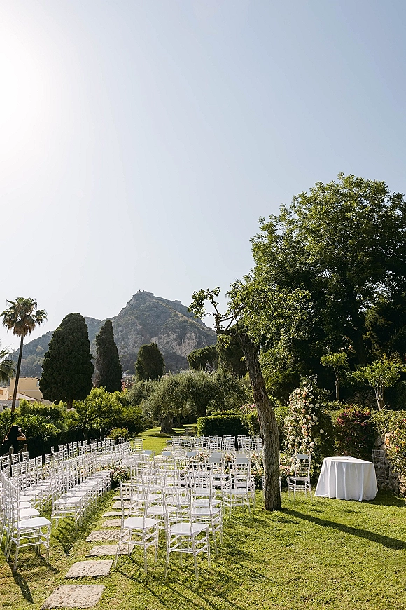 Outdoor ceremony setup with white chiavari chairs facing a floral arch along a curved stone aisle runner on a garden lawn with mountain views