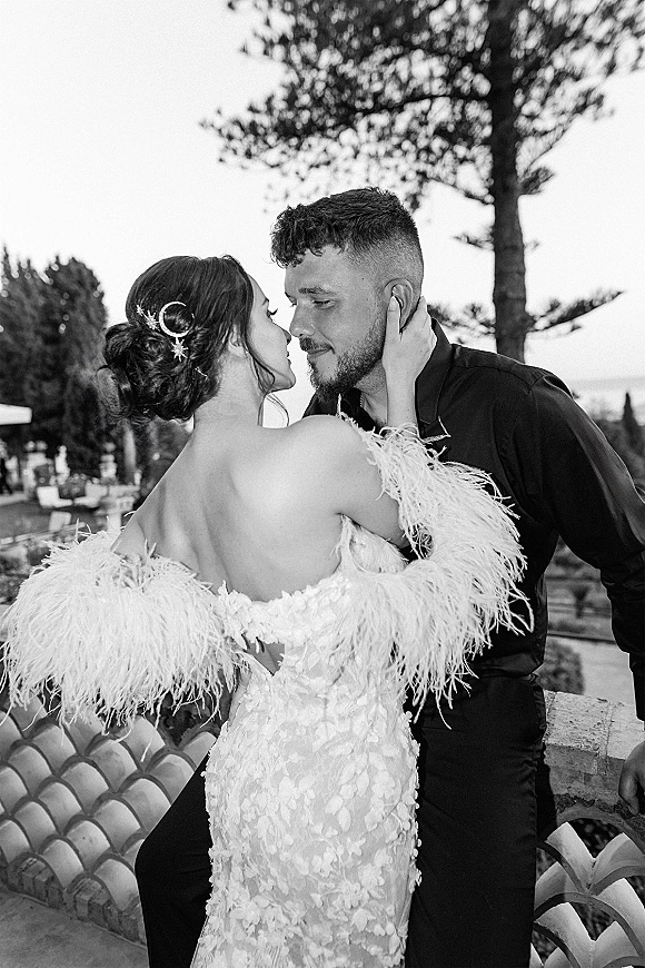 Wedding couple portrait in black and white, bride in lace gown with feather sleeves touching groom’s face nose to nose on a garden terrace by water