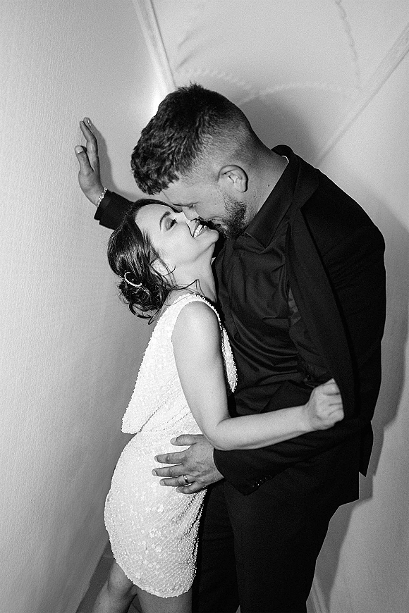 Wedding couple portrait with a wedding kiss portrait as the groom hugs the laughing bride by an indoor wall near a staircase, her hair clip visible