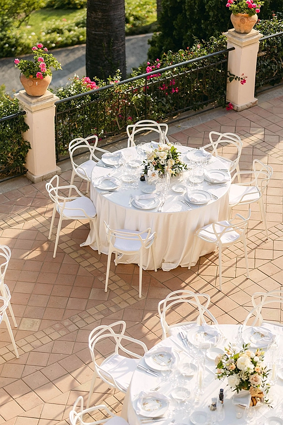 Reception tablescape at an outdoor wedding reception with white tablecloth, pink floral centerpieces, glassware, and terracotta planters on a garden patio