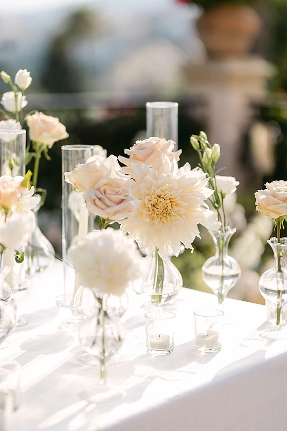 Wedding tablescape with a white wedding centerpiece of bud vases, blush roses, and votive candles on linen in a garden setting
