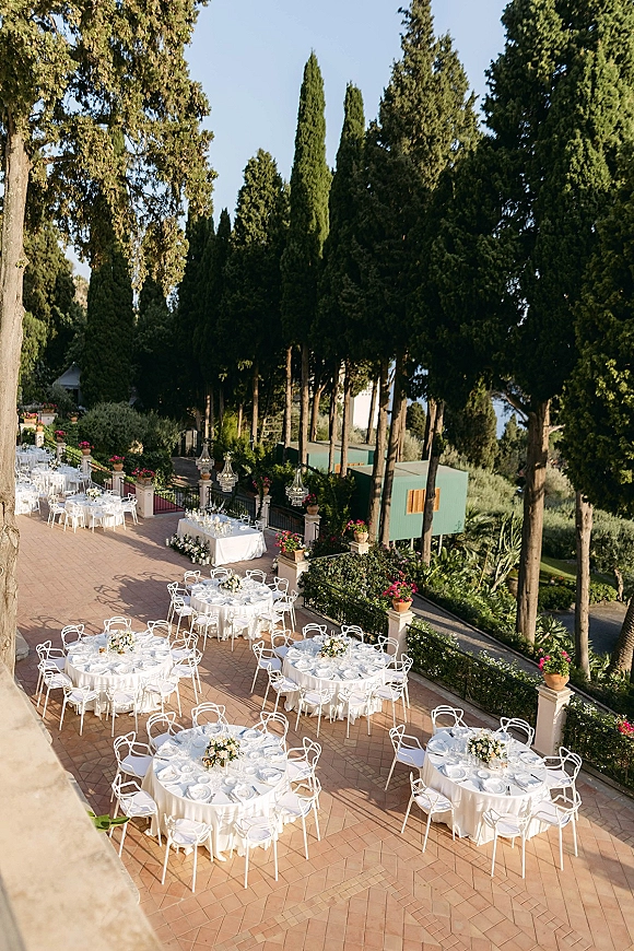 Outdoor reception setup with round dining tables, white tablecloths and floral centerpieces beneath chandeliers on a garden terrace patio