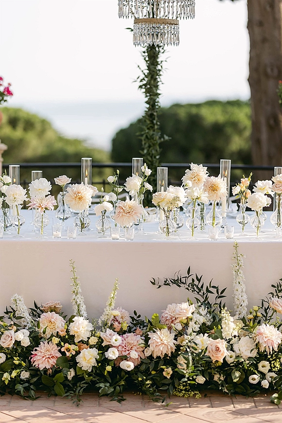 Reception sweetheart table with sweetheart table decor of blush and white florals, bud vases, greenery garland, and candlelight under a crystal chandelier on a terrace with water view