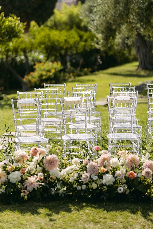 Ceremony seating with clear chiavari chairs arranged in rows, lined by blush and white floral aisle arrangements on a sunny garden lawn