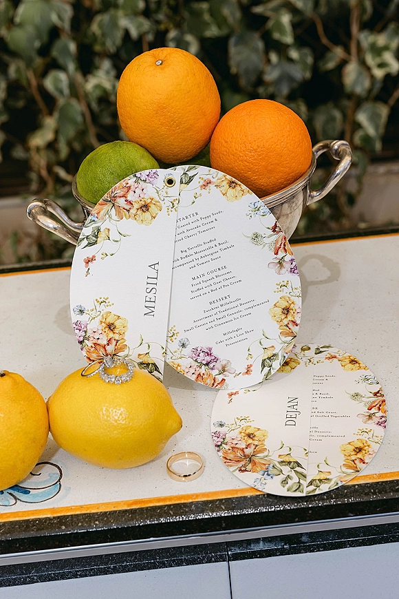 Wedding menu cards with round wedding menu design and floral accents, styled with citrus in a silver bowl and rings on a countertop with greenery