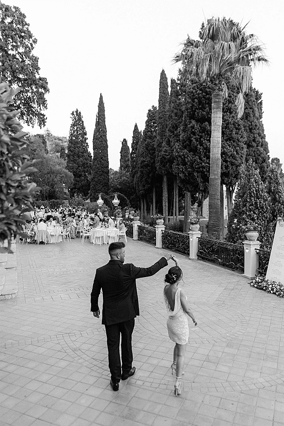 Couple dancing at an outdoor wedding reception, bride in a backless dress and heels with groom in tux on a terrace patio with palms