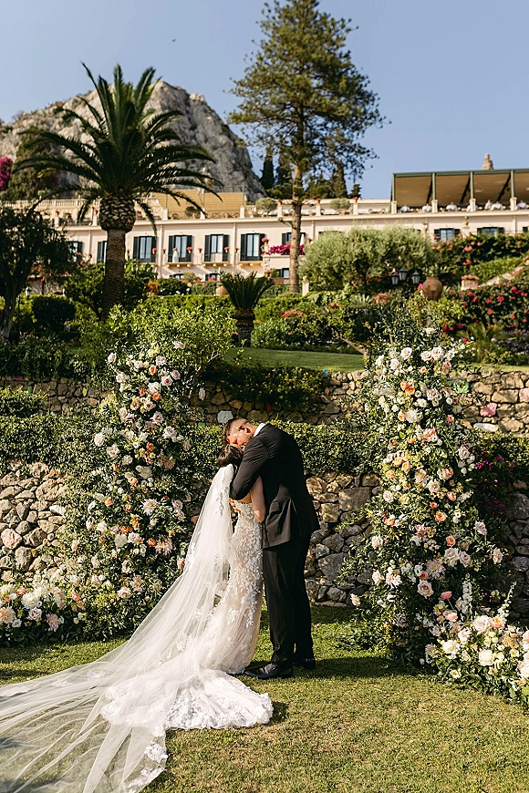 Wedding couple portrait of bride and groom embrace beneath a floral arch, lace gown and long veil against a stone wall and blue sky