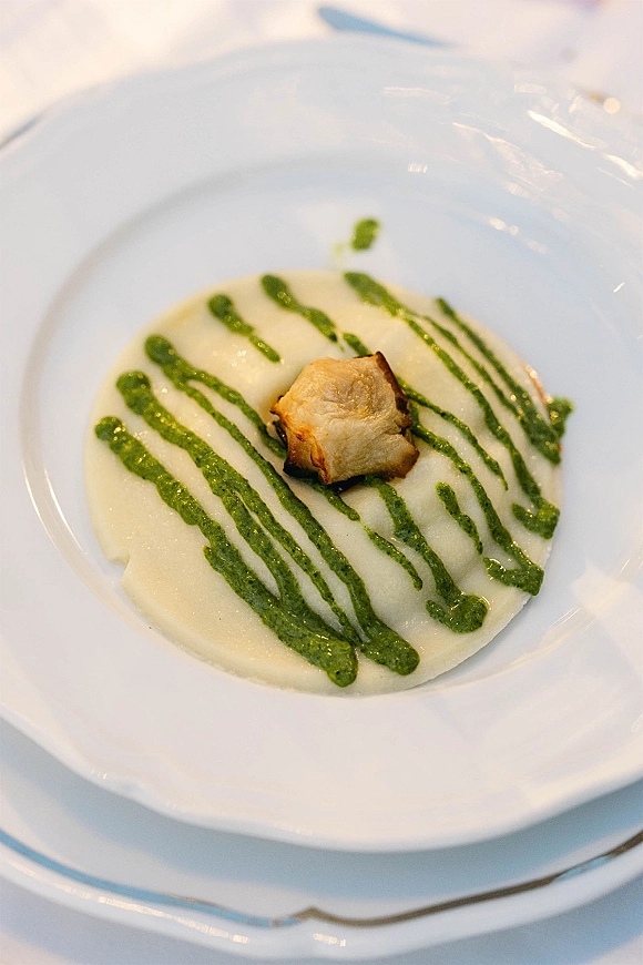 Plated wedding appetizer served as a wedding starter course on a white dinner plate with puree base, herb sauce drizzle, and roasted garnish on a white tablecloth