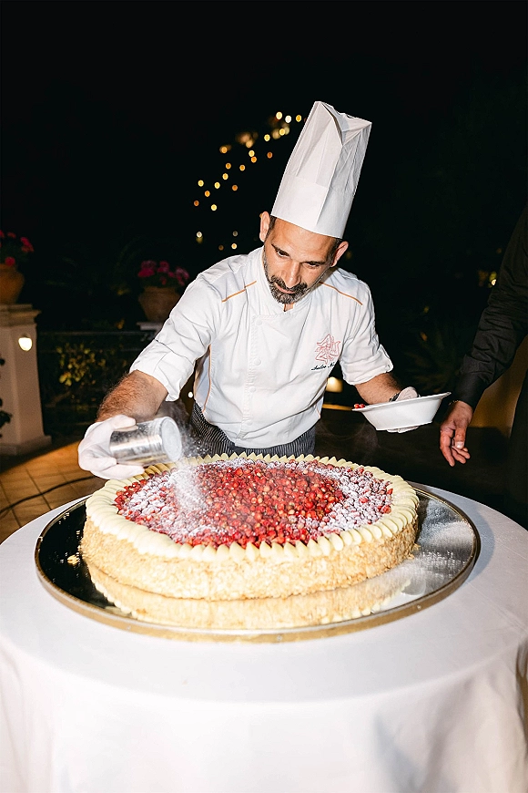 Wedding cake with berry topped wedding cake and powdered sugar as a chef in hat and gloves plates it on a patio under string lights at night