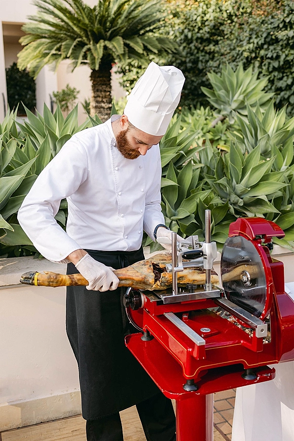 Wedding catering chef carving station with a gloved chef slicing a cured ham leg at a patio table amid tropical greenery and palms