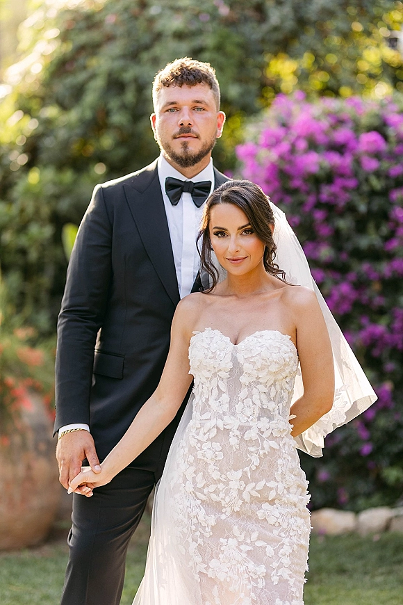 Couple portrait of bride and groom holding hands, her strapless lace dress and veil beside his black tuxedo in a garden with purple blooms