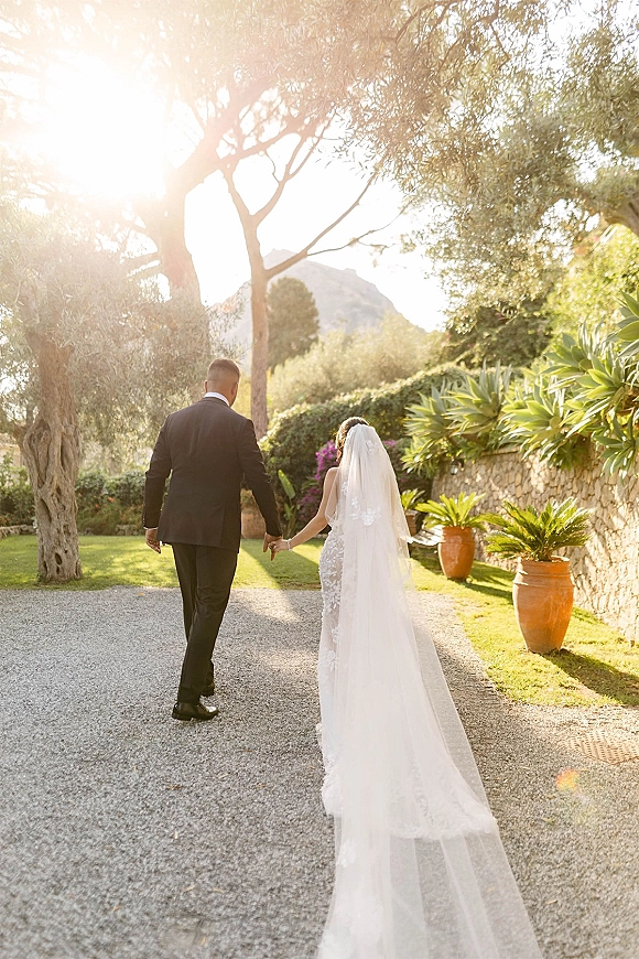 Couple walking away holding hands, bride’s long veil over lace dress and groom in black suit on a sunlit garden path by stone wall and mountain backdrop