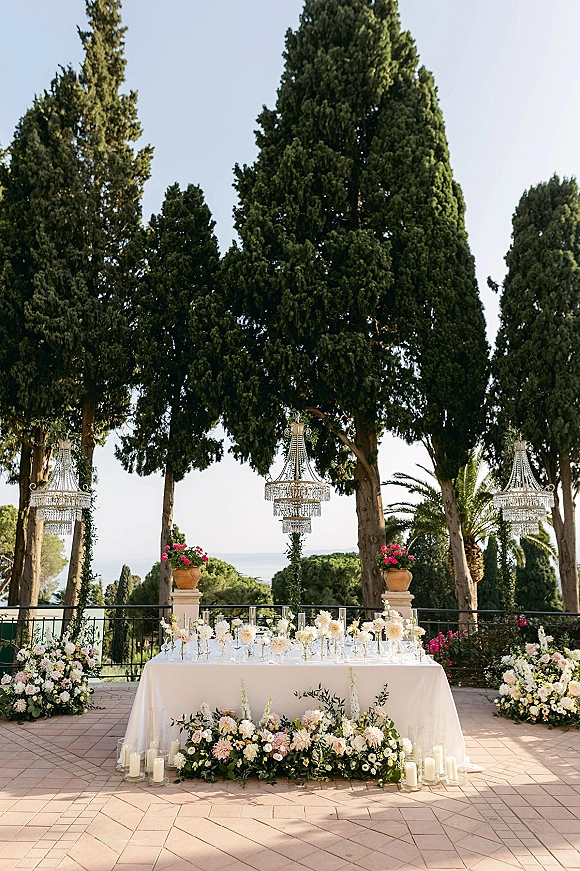 Sweetheart table decor with an outdoor sweetheart table set in white linen, lush floral garland and candles on a sea-view terrace under chandeliers