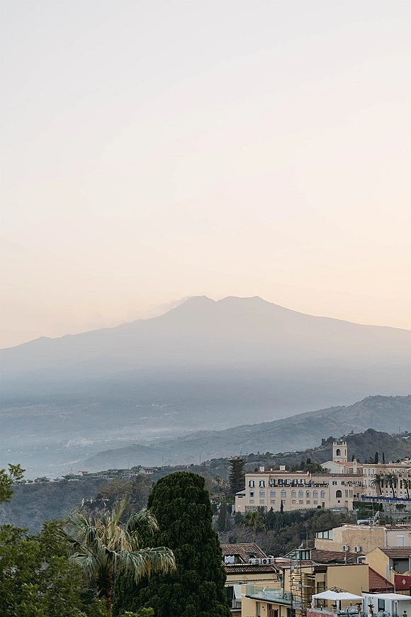 Mountain landscape with misty mountain view above a hillside town, palm trees and a church tower silhouetted under a pastel dusk sky