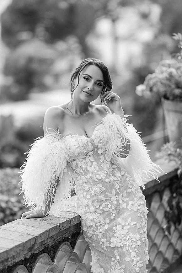 Bridal portrait in black and white of a bride in a strapless lace wedding dress, leaning on a stone balustrade with garden foliage behind