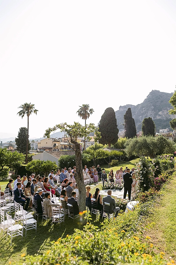 Outdoor wedding ceremony with bride and groom at a floral arch, guests in white chairs on a lawn with palm trees and mountains behind