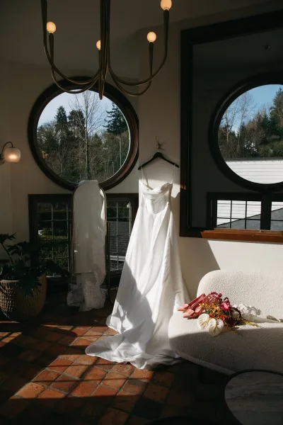 Wedding dress hanging by a round window in daylight, with lace-trim veil, pink bridal shoes, and anemone bouquet on brick floor