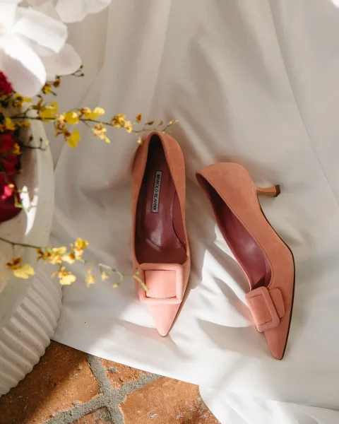 Wedding shoes in a bridal shoes flat lay with pointed-toe buckle heels beside a flower vase on white fabric, brick floor, and sunlit shadows