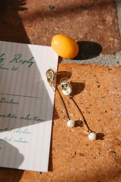 Wedding jewelry flatlay with gold pearl drop earrings on pink striped stationery and RSVP card, set on terracotta tile in hard sunlight shadows