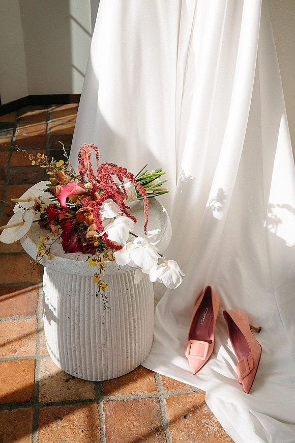 Wedding detail flatlay featuring a bridal bouquet and shoes beside a wedding dress on a pedestal table, lit by window light on a brick floor