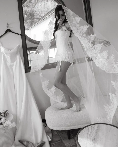 Bridal portrait of a bride in a lace veil and corset lingerie by an arched window in an indoor suite, with bouquet and dress on hanger nearby