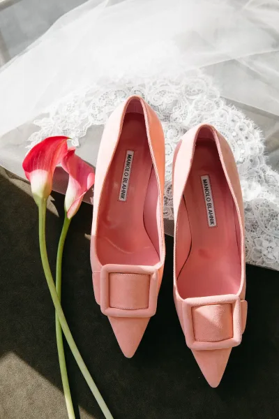 Wedding shoes in blush pink heels with square buckle detail beside calla lilies and a lace veil on gray fabric with sunlit shadows