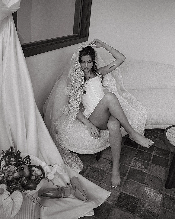 Bridal portrait of a bride in corset lingerie adjusting a lace veil on a couch, with bouquet and framed mirror on tiled floor